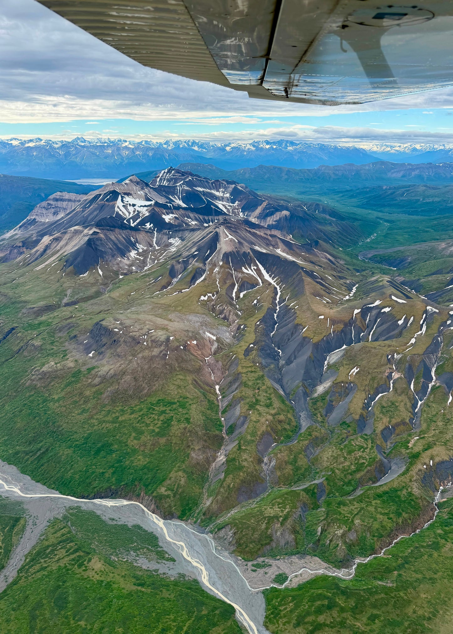 Aerial view of rugged mountain peaks with patches of snow, green valley terrain, and a winding river below, with part of an airplane wing visible at the top edge.