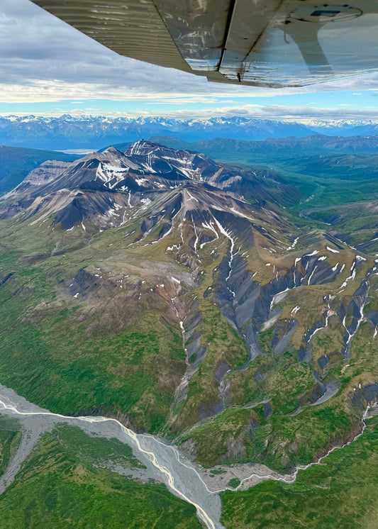 Aerial view of rugged mountain peaks with patches of snow, green valley terrain, and a winding river below, with part of an airplane wing visible at the top edge.